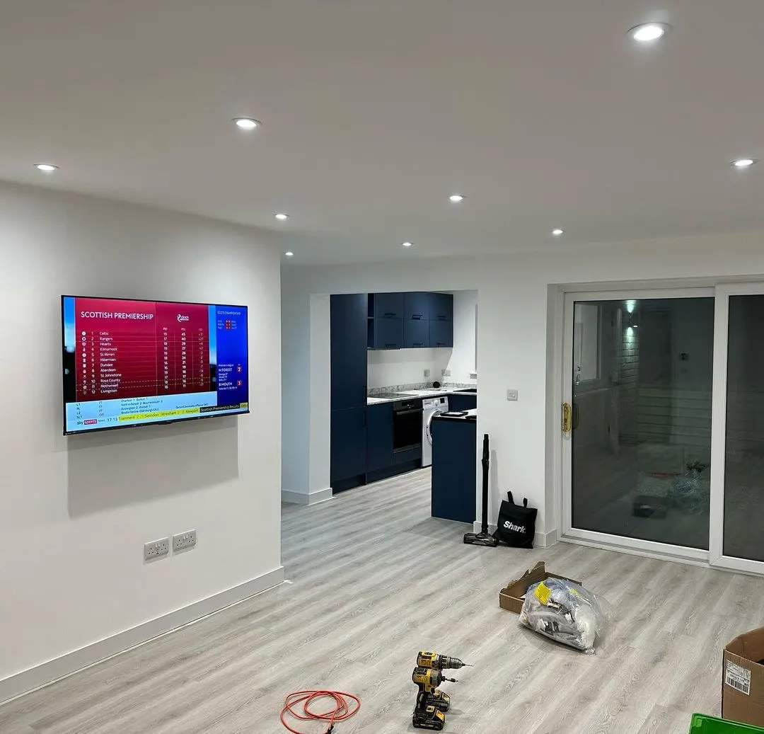 Open-plan room with fitted downlights, wall television, kitchen beyond and tools on the floor