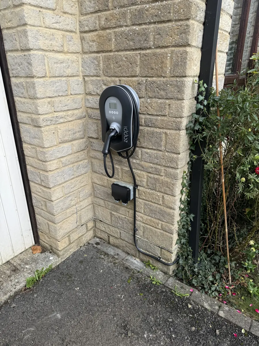 Black zappi charger with tethered cable on a stone wall, with a small black enclosure below and plants beside it