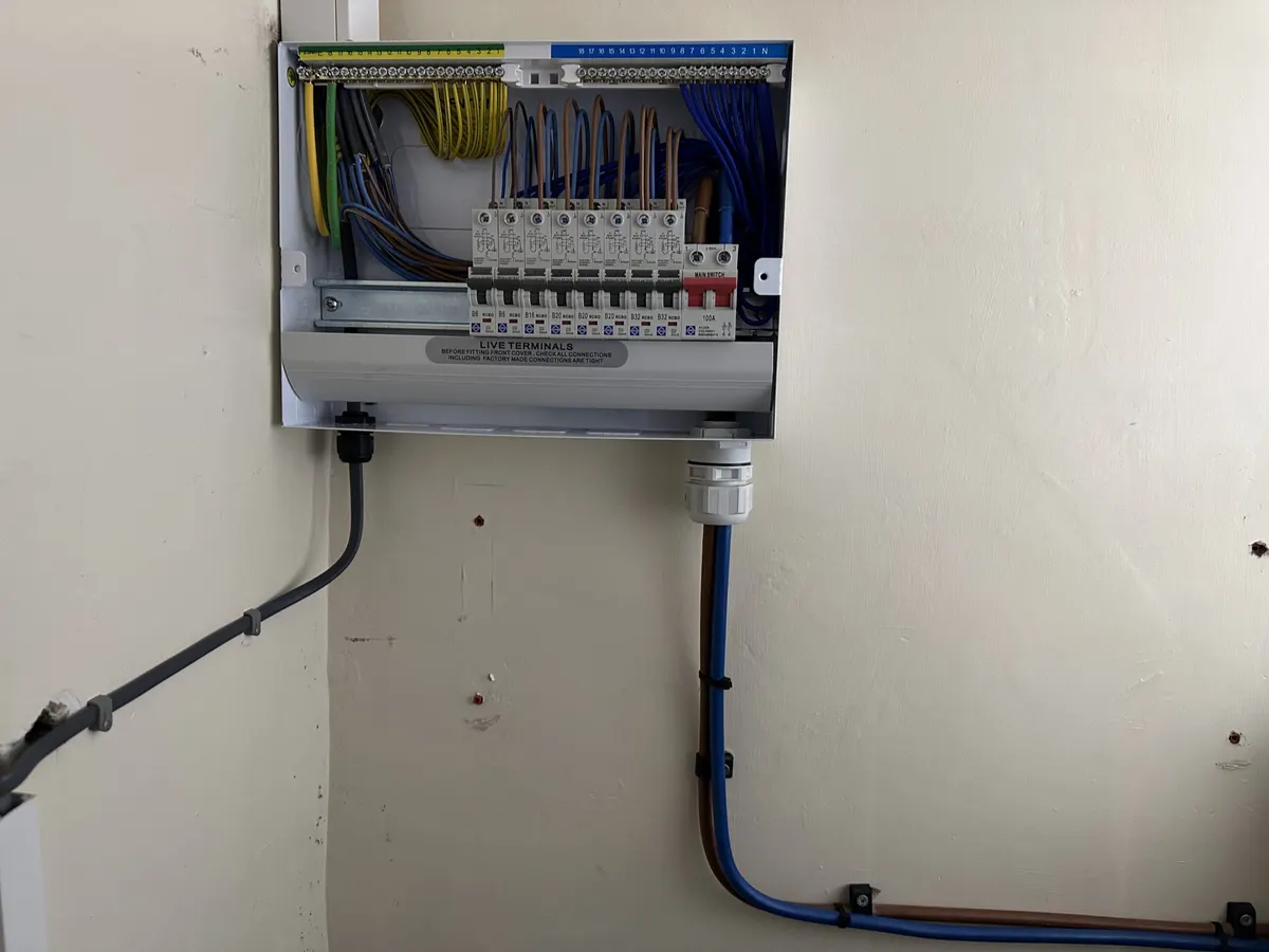 Consumer unit with front cover removed showing wiring and circuit devices, with a blue supply cable entering below