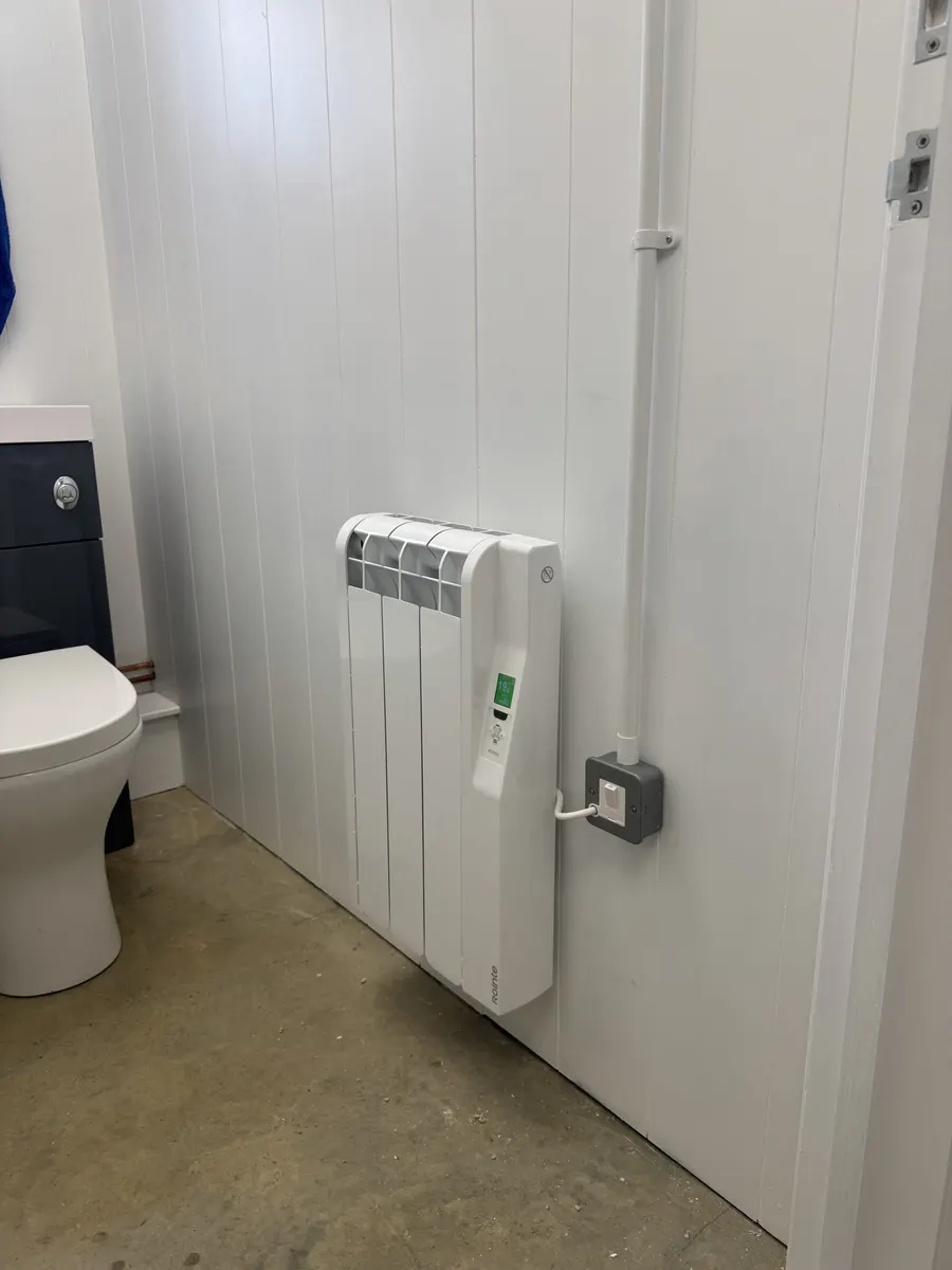 White electric radiator beside a toilet with a grey switched connection plate and vertical white conduit