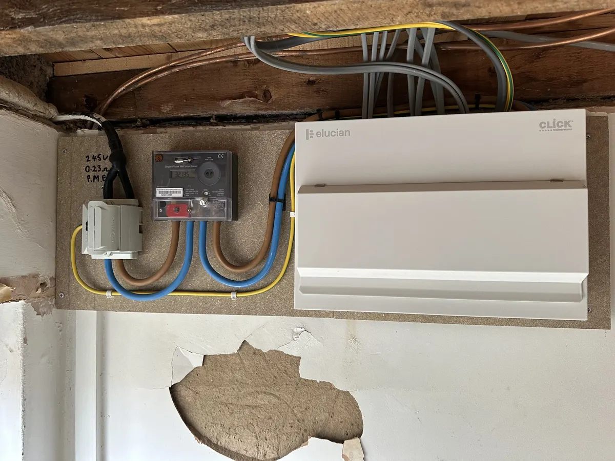 Older cabling and a junction box on a wall beside a newer white electrical enclosure, with patched plaster below
