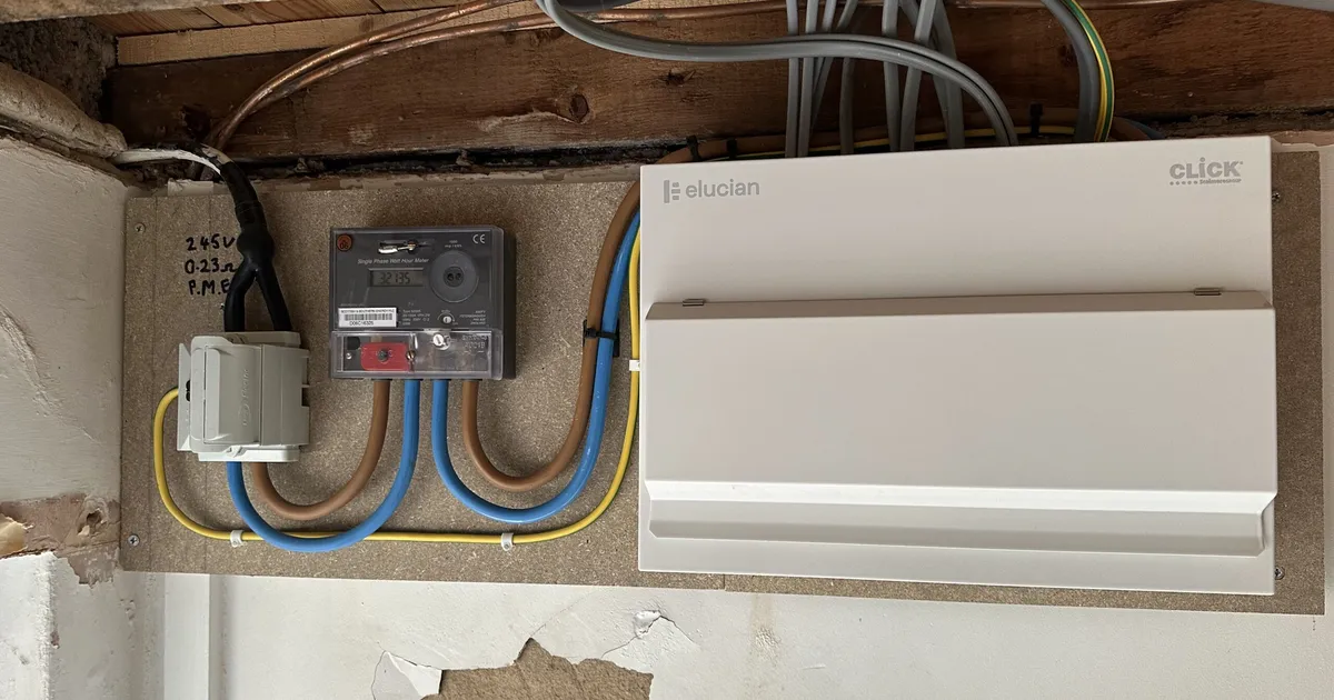 Older cabling and a junction box on a wall beside a newer white electrical enclosure, with patched plaster below