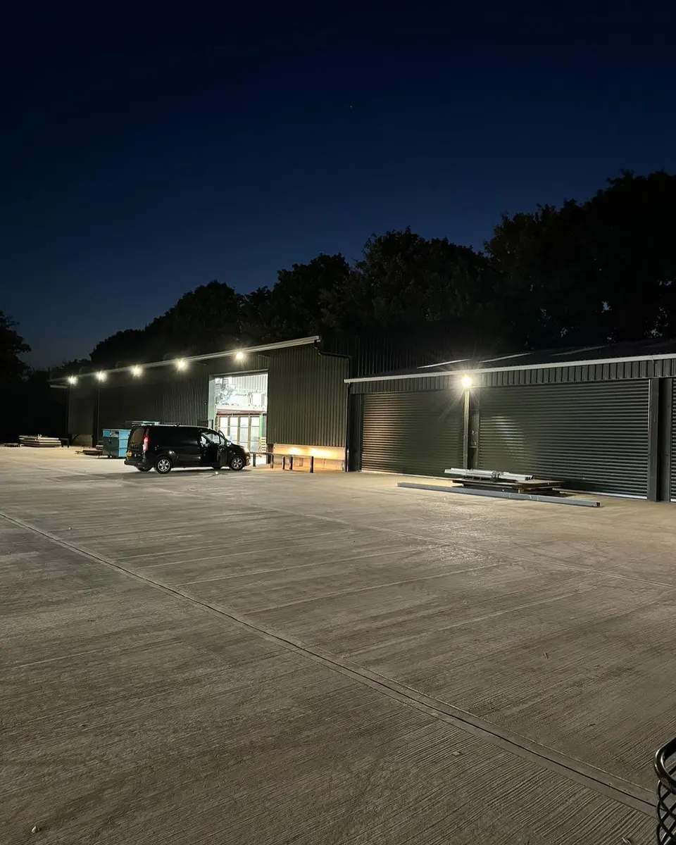 Commercial buildings and concrete yard at night, with wall-mounted floodlights and a dark van beside an open doorway