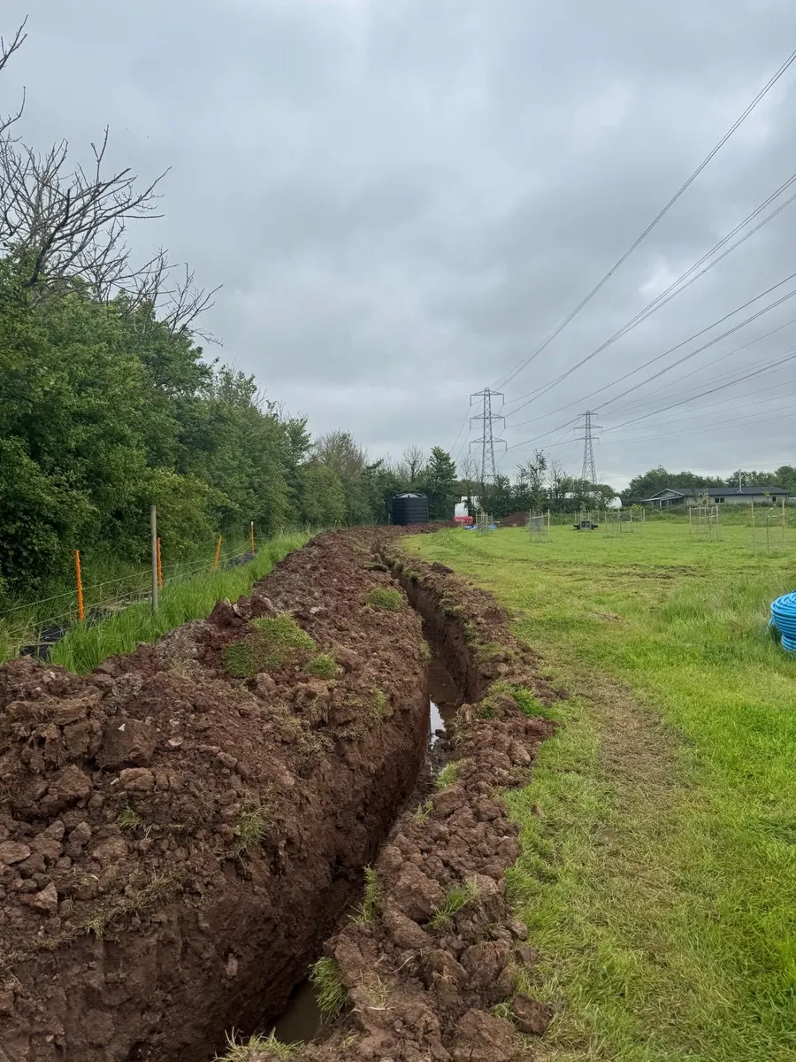 Long cable trench cut across a field beside a hedge, water tank and pylons in the distance
