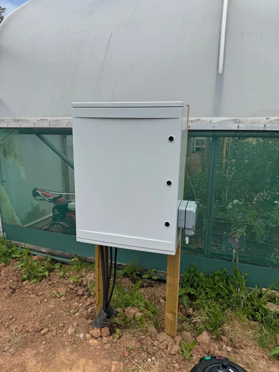 Closed grey outdoor enclosure on timber posts beside a polytunnel, cables below and a weatherproof socket on the side